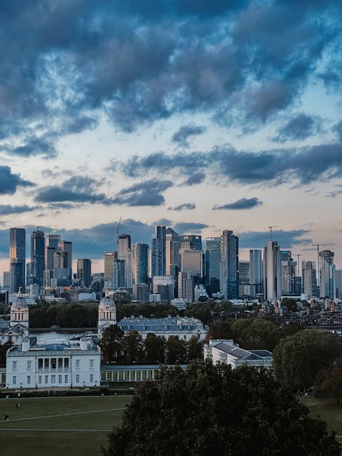 A wide panoramic view of a cityscape with a large, historic white building situated in a spacious, well-maintained green park in the foreground. The building features classical architectural elements,