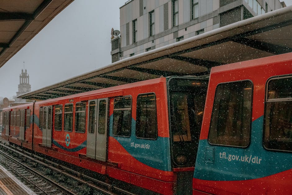 A modern red and blue light rail train is stationed at an outdoor platform in an urban setting, with snowflakes gently falling around it. The train's exterior features a smooth, metallic finish with l