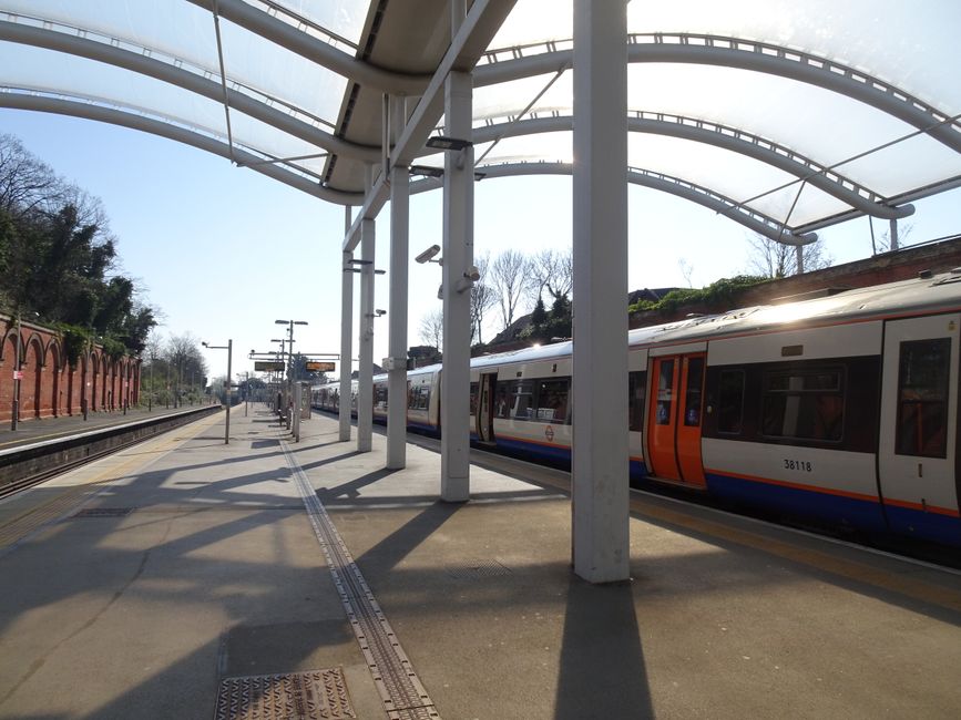 A modern red and blue light rail train is stationed at an outdoor platform in an urban setting, with snowflakes gently falling around it. The train's exterior features a smooth, metallic finish with l